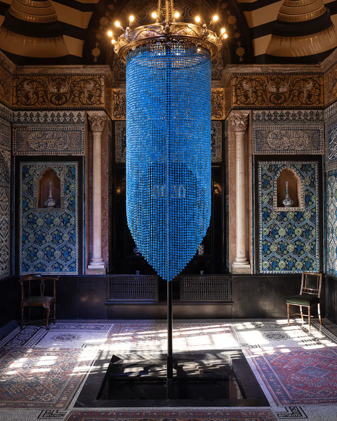 Ornate Islamic interior with large blue beaded chandelier, intricate geometric tilework, Arabic calligraphy panels, arched niches, vintage chairs, patterned carpet, and sunlight casting shadows across a historic palace or mosque hall.